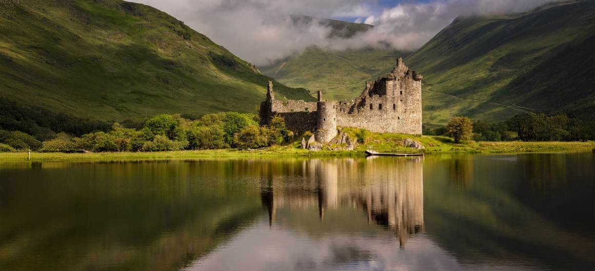 Die Ruinen von Kilchurn Castle spiegeln sich im ruhigen Wasser des Loch Awe, umgeben von nebligen grünen Hügeln in Argyll, Schottland.