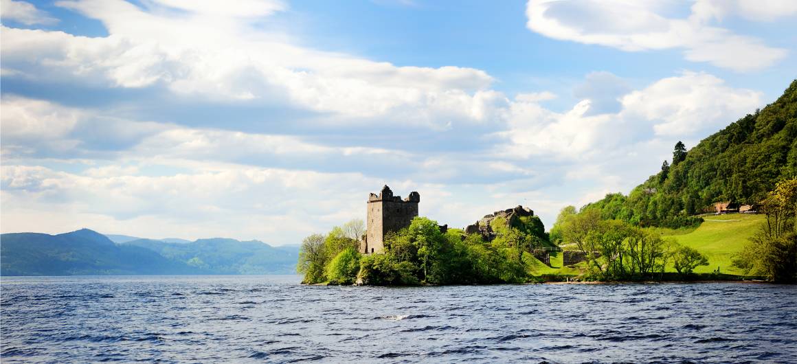 Eine teilweise erhaltene Steinburg liegt am Ufer des Loch Ness, umgeben von üppig grünen Bäumen und grasbewachsenen Hügeln.