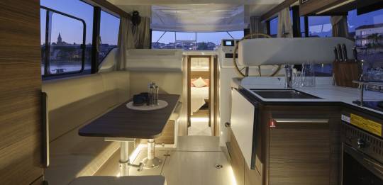 Interior of the Liberty boat at dusk featuring a saloon with a dining area, helm station, and a view into the bedroom with warm ambient lighting.