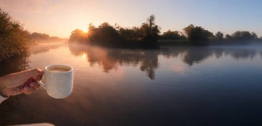 A hand holding a cup of coffee while watching the sunrise over a quiet river during a relaxing boating escape.
