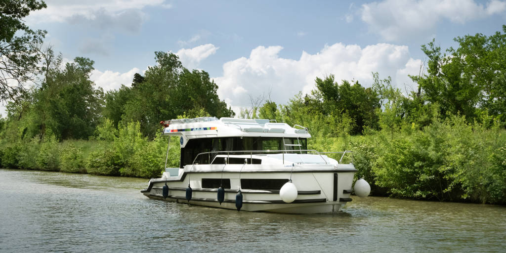 Liberty boat gliding gently on the water, with lush foliage along the banks and soft summer skies above.
