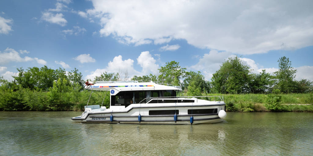 Das Boot Liberty beim Gleiten entlang eines grün bewachsenen Flussufers, mit elegantem Profil und schattigem Flybridge-Bereich – gespiegelt im stillen Wasser.
