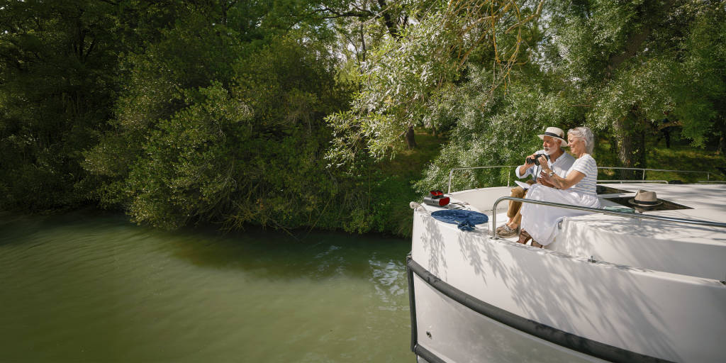 Couple birdwatching together from the front of their canal boat, surrounded by lush green forest.