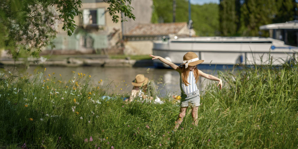 Young girl playing in the grass near the river, with a moored boat in the background on a summer day.