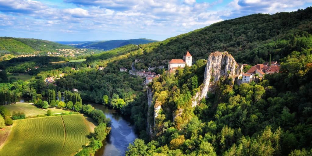 Scenic view of the Lot River at the base of tall limestone cliffs, surrounded by lush greenery.