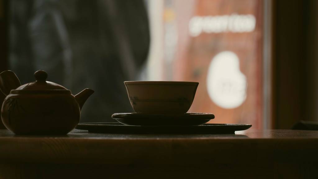 Silhouetted clay teapot and tea bowl on saucers atop a wooden table, with a blurred "green tea" sign in the background.