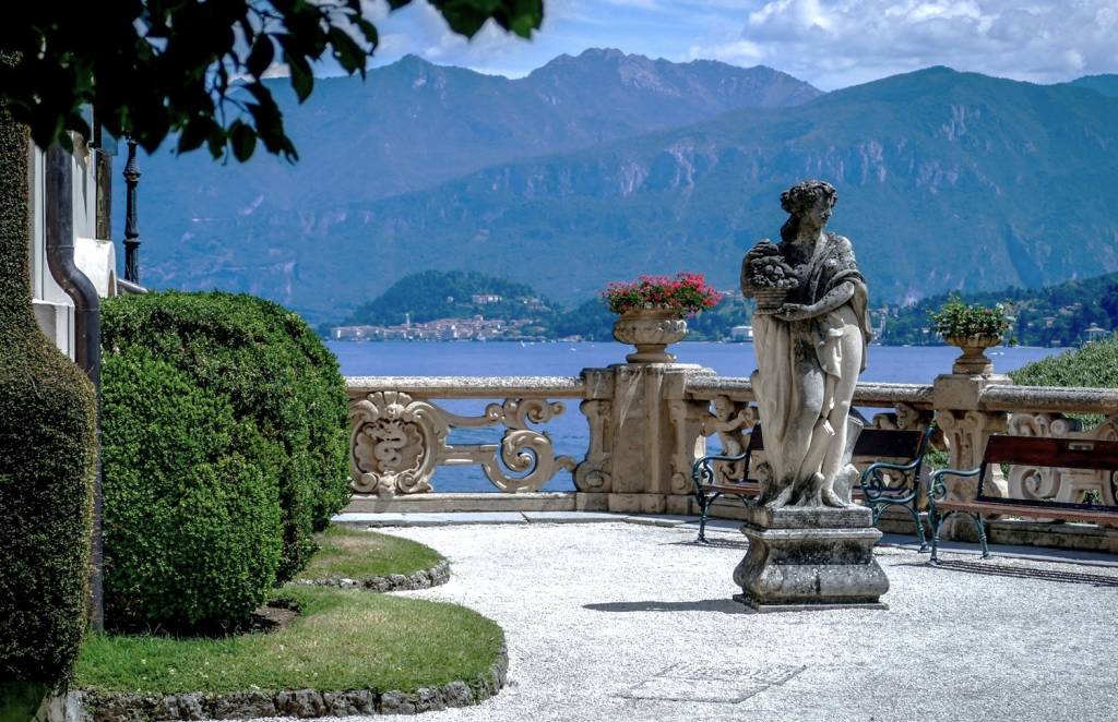 Classical stone statue on an ornate terrace overlooking Lake Como, Italy, with mountains and a village in the background.