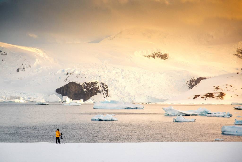 Panoramablick auf Eisberge und stille See im goldenen Abendlicht. Im Vordergrund ein einsamer Fotograf mit Stativ.