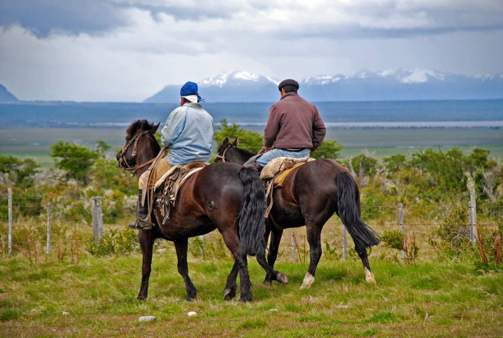 Rückansicht zweier Gauchos auf dem Rücken der Pferde in Patagonien, im Hintergrund schneebedeckte Berge