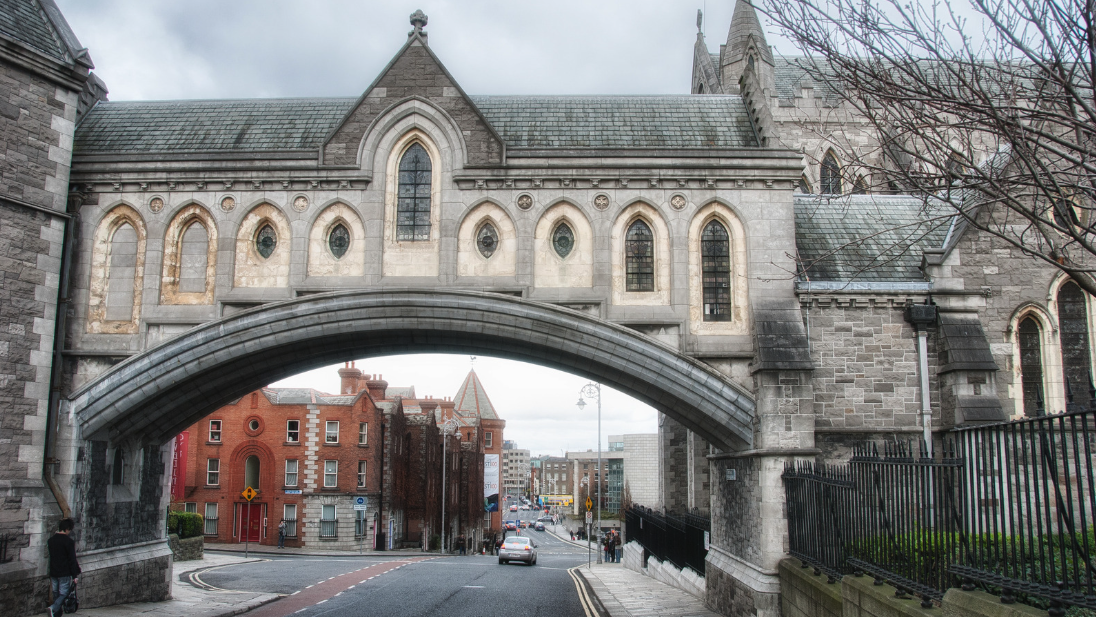 The iconic **synod hall bridge of Christ Church Cathedral in Dublin, Ireland**. A Gothic stone archway bridge connects the medieval cathedral (right) to the synod hall building (left) over a city street below. The bridge features ornate Gothic-style arched windows and stonework. Through the arch, a street scene is visible with red-brick Victorian buildings, parked cars, and pedestrians. A bare tree stands to the right, and iron railings line the sidewalk. The sky is overcast and cloudy.