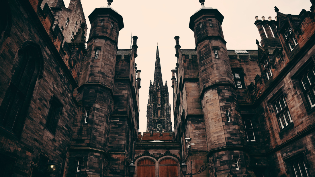 A low-angle view of a grand Gothic-style stone building complex, likely in Edinburgh, Scotland. The foreground features a symmetrical facade with two octagonal corner towers flanking a central arched wooden gate. Behind it rises a tall, ornate Gothic church spire with a clock, framed between the towers against an overcast sky. The dark reddish-brown sandstone architecture creates a dramatic, moody atmosphere.