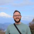 Smiling bearded man with glasses and crossbody bag, Mount Fuji's snow-capped peak visible in the background.