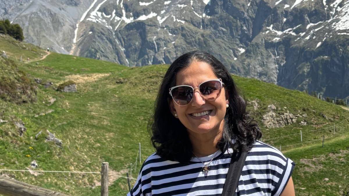 Vineeta smiling outdoors in sunglasses and a striped shirt, with snow-capped alpine mountains and green meadows behind her.