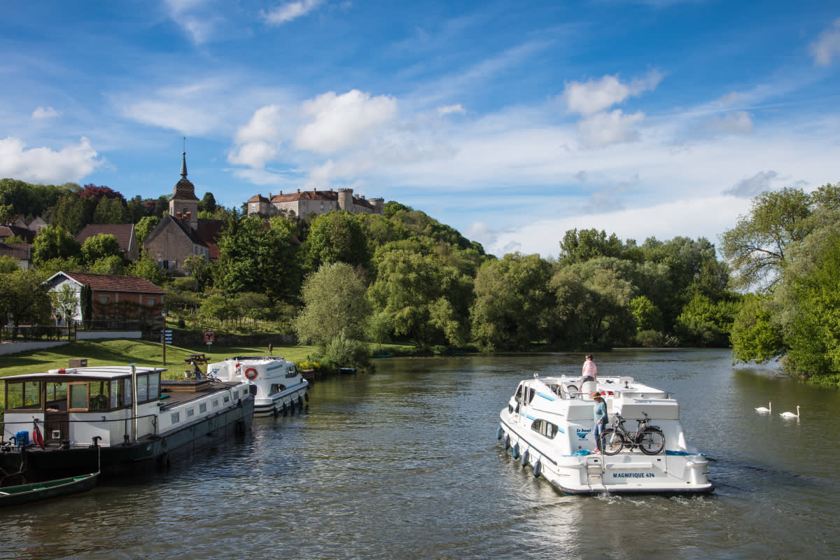 A white canal boat with a bicycle on the deck cruising along a calm river or canal, passing other moored boats. In the background, a picturesque French town with a prominent church spire and a historic castle or fort on a green hill overlooks the waterway in Burgundy-Franche-Comté, France.