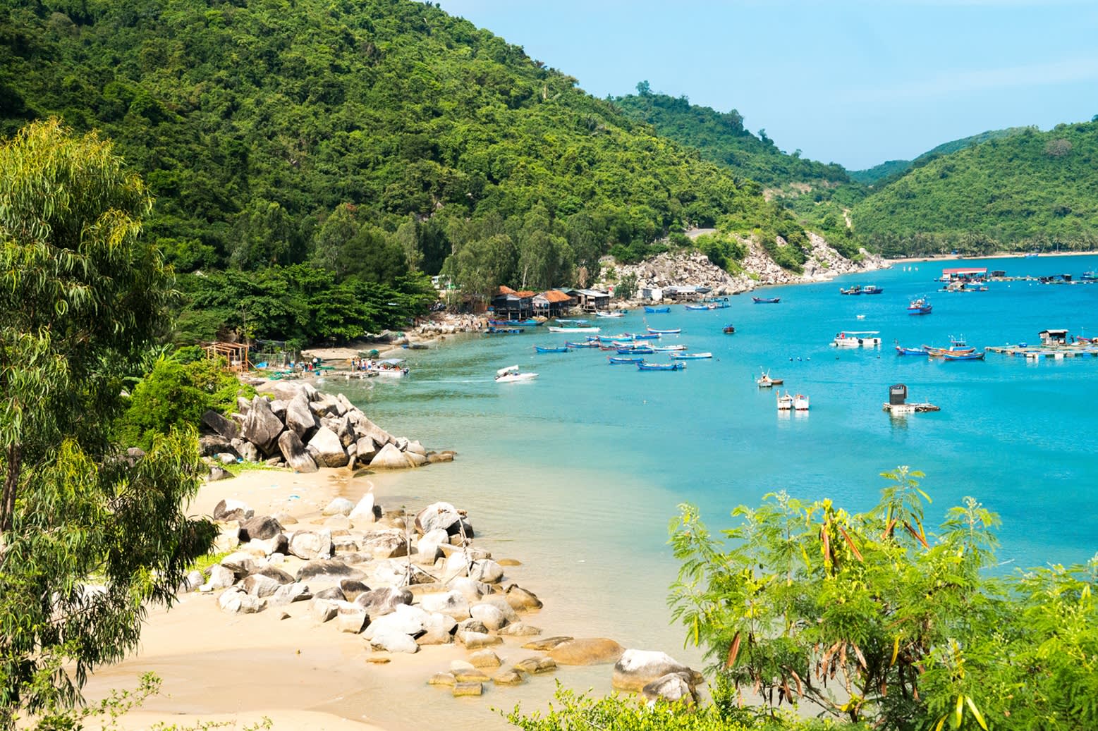 Blick auf einen schmalen Sandstrand  in Quy Nhon, Vietnam mit tropisch bewachsenen Hügeln. Auf dem türkisfarbenem Meer ankern kleine Holzboote. 