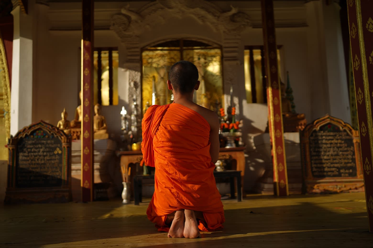 Rückansicht eines in eine orangefarbene Robe gekleideten, in einem Tempel betenden thailändischen Mönches. 