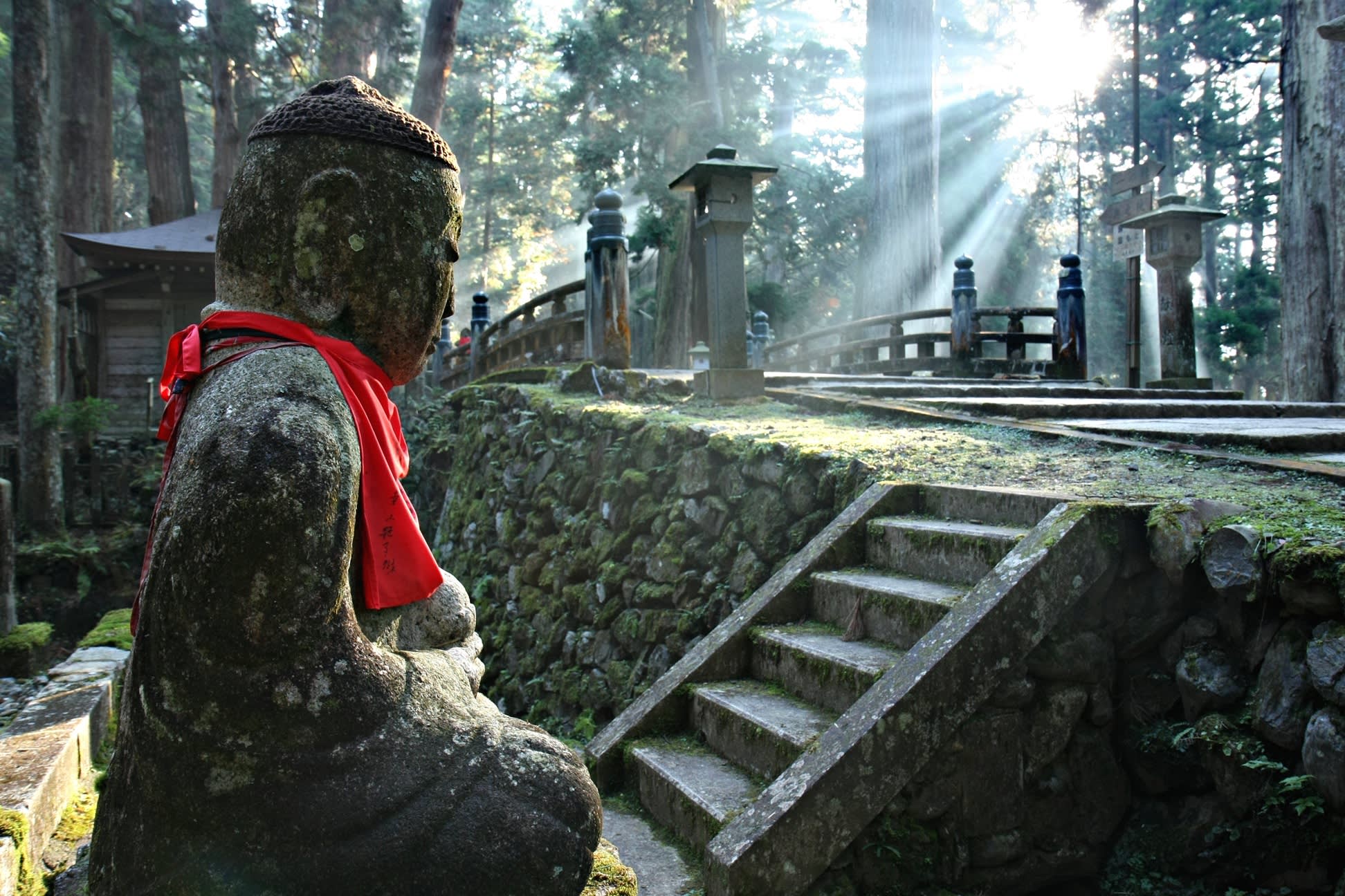 Moosbedeckte Buddha-Statue mit rotem Schal in Koyasan, Japan, umgeben von Treppen, Steinlaternen und Sonnenstrahlen im Wald.