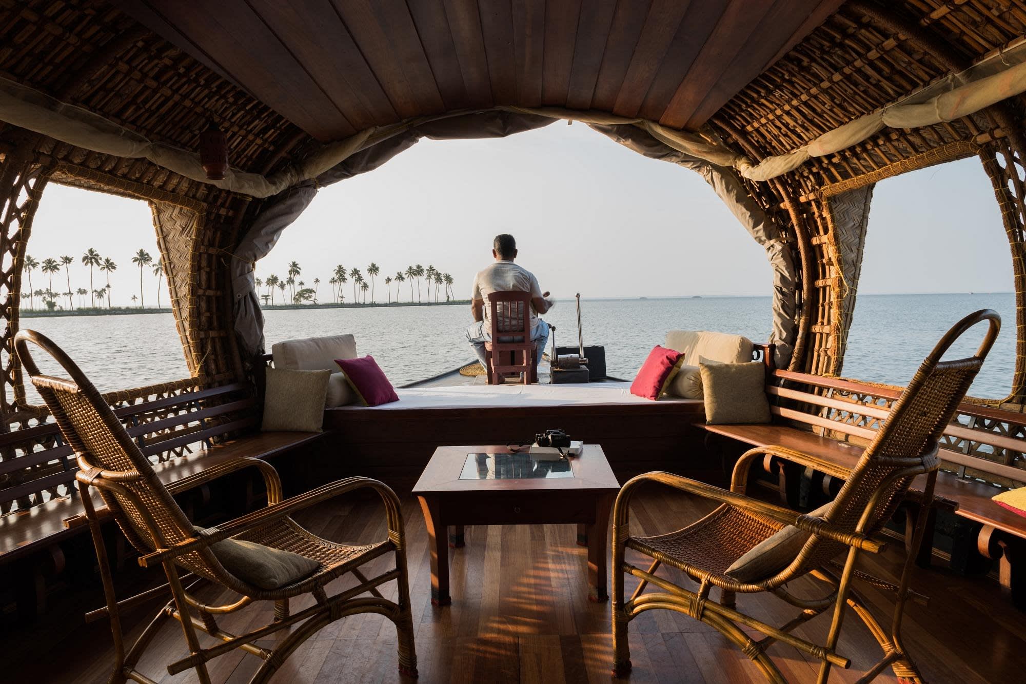 Blick aus einem Hausboot auf eine Sitzgruppe, den Bootsführer und Wasserlandschaft der Kerala Backwaters