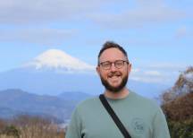 Smiling bearded man with glasses and crossbody bag, Mount Fuji's snow-capped peak visible in the background.