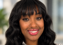 Bre Epps smiling, with long wavy black hair with bangs, nose ring, silver necklace, and patterned white blouse.