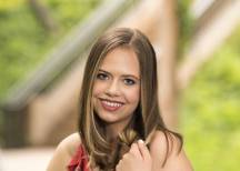 Young woman with long brown hair smiling, wearing a red floral top and charm bracelet, outdoors with blurred greenery.