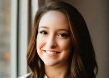 Rachel Cronin, a young woman with long wavy brown hair, smiling warmly, wearing a white button-up shirt near a window.