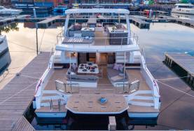 Power catamaran stern showing a swim platform, aft cockpit with seating and a dining table, and a flybridge with helm and lounge seating above.