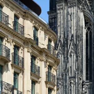 Renovated façade of one of Europe's oldest grand hotels, featuring a cream-colored Beaux-Arts or Neo-Classical building with ornate wrought-iron balconies across multiple floors and a curved corner section. Adjacent to the right stands the dark stone Gothic architecture of what appears to be Cologne Cathedral, with its characteristic pointed arches, flying buttresses, and intricate stone tracery. The contrast between the elegant hotel's light limestone exterior and the cathedral's dark Gothic stonework is striking under a clear blue sky.