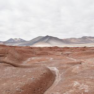 Karge, rotbraune Felslandschaft der Atacama-Wüste in Chile. Im Vordergrund erstreckt sich eine erodierte, wellige Gesteinsformation, im Hintergrund sind dunkle Vulkankegel sowie ein heller Salzsee (Salar) unter bewölktem Himmel zu sehen.