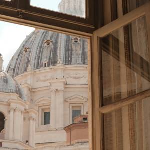 View through an open wooden window of St. Peter's Basilica's dome in Vatican City, with ornate carved wall detail visible.