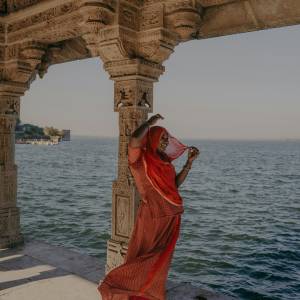 Woman in red saree posing beside ornate carved stone columns of an Indian temple overlooking a lake.