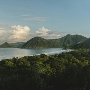 Lush tropical coastline with green forested hills, a curved bay, and dramatic clouds over mountain peaks.