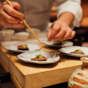 Chef carefully plating elegant small dishes with chopsticks on a wooden board in a fine dining kitchen.