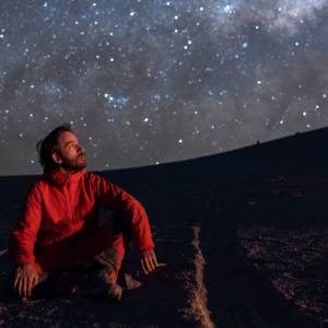 Junger Mann, auf dem Boden kniend, den Blick auf den Sternenhimmel gerichtet, in der Atacama-Wüste Chile