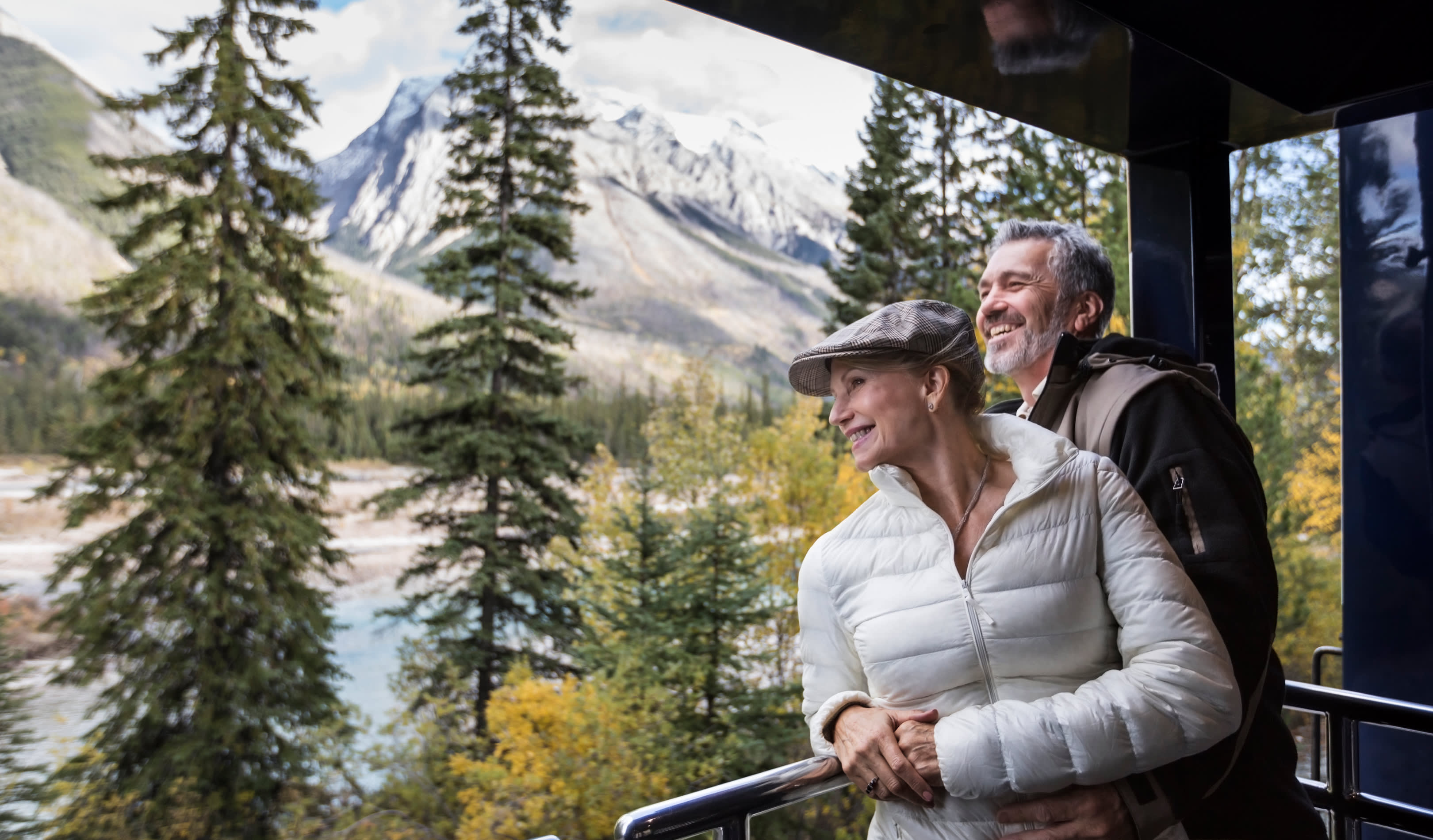 Nahaufnahme eines reisenden Paares auf der offenen Aussichtsplattform des Rocky Mountaineer Zuges in Kanada. Im Hintergrund Tannen und schneebedeckte Berge