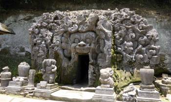 a stone statue in front of a large rock with Goa Gajah in the background