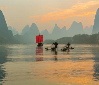 Fisherman stands on traditional bamboo boats at sunrise (boat with a red sail in the background)