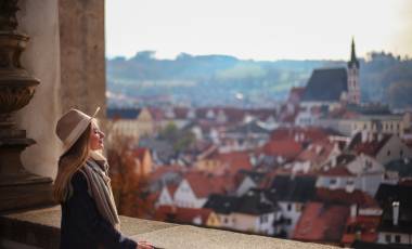 A young woman wearing a beige wide-brimmed hat, scarf, and dark coat stands at a stone balcony railing, eyes closed and facing the sun. Behind her stretches a panoramic view of Český Krumlov, Czech Republic, featuring a dense cluster of red-roofed buildings, a prominent church with a steeple, and rolling green hills in the background.