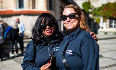 Two smiling women wearing matching navy blue "Europe Express" zip-up jackets and sunglasses pose together outdoors on a sunny day during the AAT Croatia & Slovenia 2025 trip. One woman has her arm around the other's shoulder. A stone plaza and historic building are visible in the blurred background.