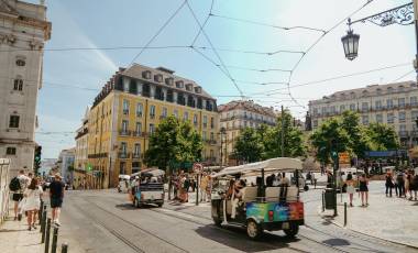 A busy urban square in Lisbon, Portugal, featuring tuk-tuks (small motorized tourist vehicles) navigating cobblestone streets with tram tracks. The scene is framed by classic European architecture, including a prominent yellow building with mansard roof, surrounded by green trees. Overhead tram wires crisscross above the intersection, and a decorative iron street lamp is visible in the upper right. Pedestrians walk along the sidewalks, and a statue is partially visible among the trees in the background. The sky is bright blue with light clouds.
