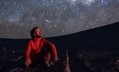 Junger Mann, auf dem Boden kniend, den Blick auf den Sternenhimmel gerichtet, in der Atacama-Wüste Chile