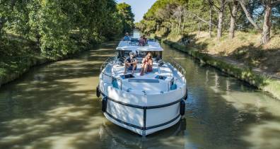 Ein Kanalboot gleitet durch einen grünen, von Bäumen gesäumten Wasserweg auf dem ruhigen Canal du Midi im Süden Frankreichs.