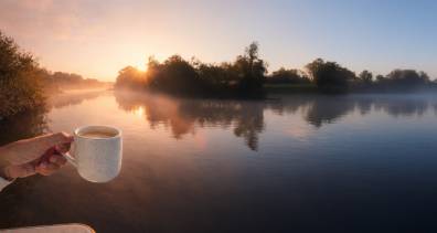 A hand holding a cup of coffee while watching the sunrise over a quiet river during a relaxing boating escape.