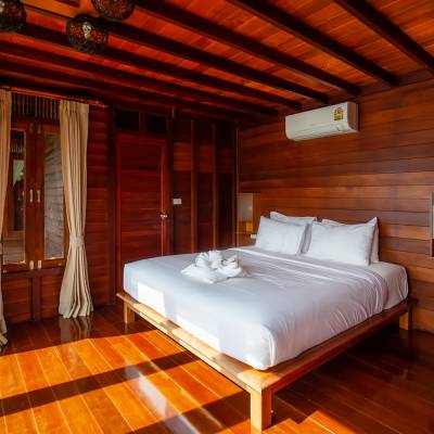 A guest room at the 9500 Rai Floating Resort Hotel, featuring a double bed with crisp white linens and towel art on a low wooden platform frame. The room has warm, rich teak wood paneling on the walls, ceiling with exposed beams, and polished hardwood floors. Amenities include a wall-mounted air conditioning unit, a small mirror, and decorative woven pendant lights. Sheer cream curtains frame glass doors leading to a balcony with a glimpse of greenery and water outside, reflecting the resort's floating nature.