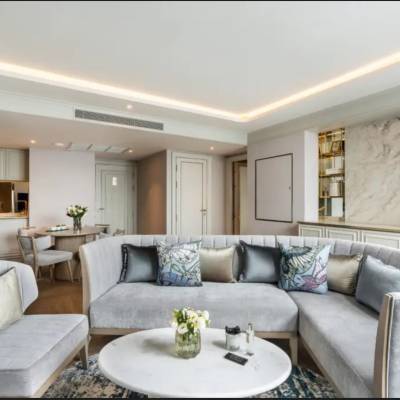 Elegant hotel suite living area with grey sectional sofa, decorative pillows, marble accent wall, and open-plan kitchen at The Salil Hotel Riverside Bangkok.