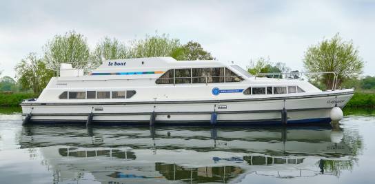 Side profile of the Classique on a quiet waterway, its white hull and upper deck mirrored on the calm canal surface.