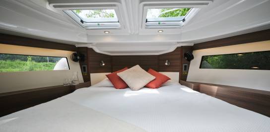 Cabine avant lumineuse à bord du Liberty, avec un grand lit, des puits de lumière au-dessus, et de grandes fenêtres panoramiques offrant une belle vue sur la rivière et la végétation.