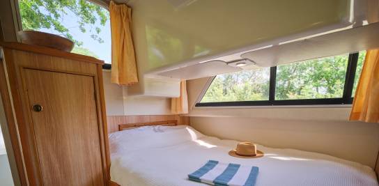 Bright single cabin inside the Cirrus B with a double bed, yellow curtains, a hat and a striped towel on the bed, and greenery visible through the side window.