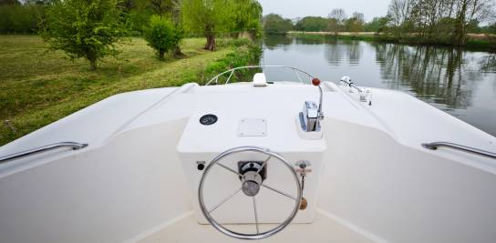 Steering position on the top deck of the Caprice, overlooking a peaceful canal with green trees and reflections lining the riverbank.