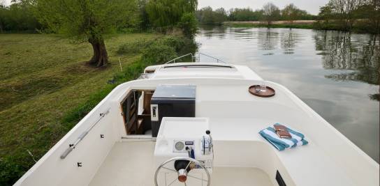 Top deck of the Classique with helm controls, a striped towel, book, and tray on the bench, overlooking a calm canal bordered by trees.
